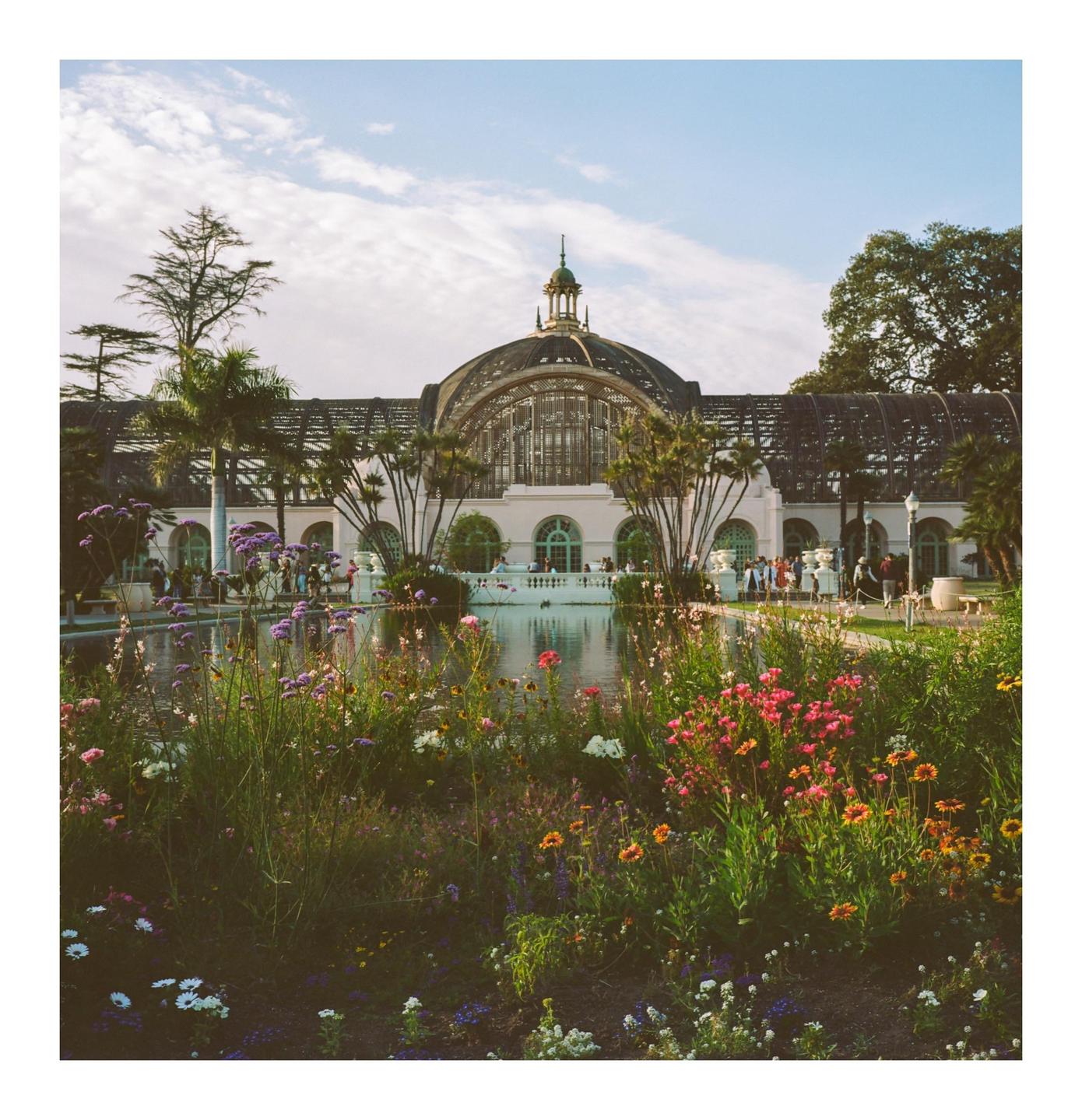 A large domed building with a pond and colorful flowers in the foreground is visible under a partly cloudy sky.