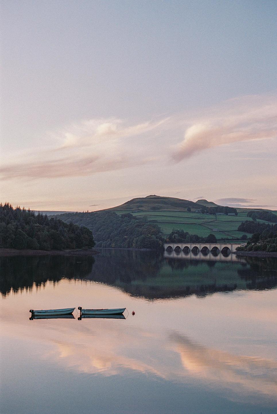 Two small boats float on a calm lake with a bridge and hills in the background under a soft sky.