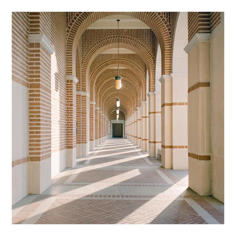 A long hallway with brick arches and columns is illuminated by sunlight creating shadows on the floor.