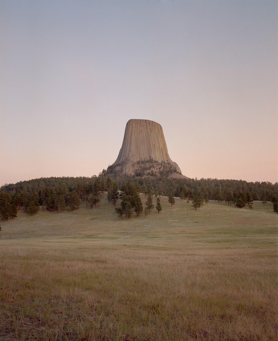 A large rock formation rises above a field and forest under a pale sky.