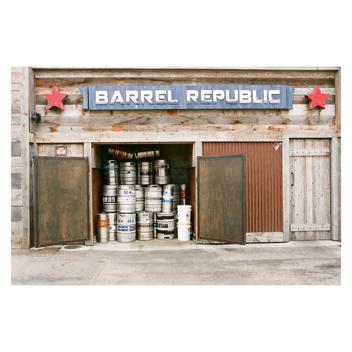 A wooden building with a sign that reads "Barrel Republic" has an open storage area filled with metal kegs.