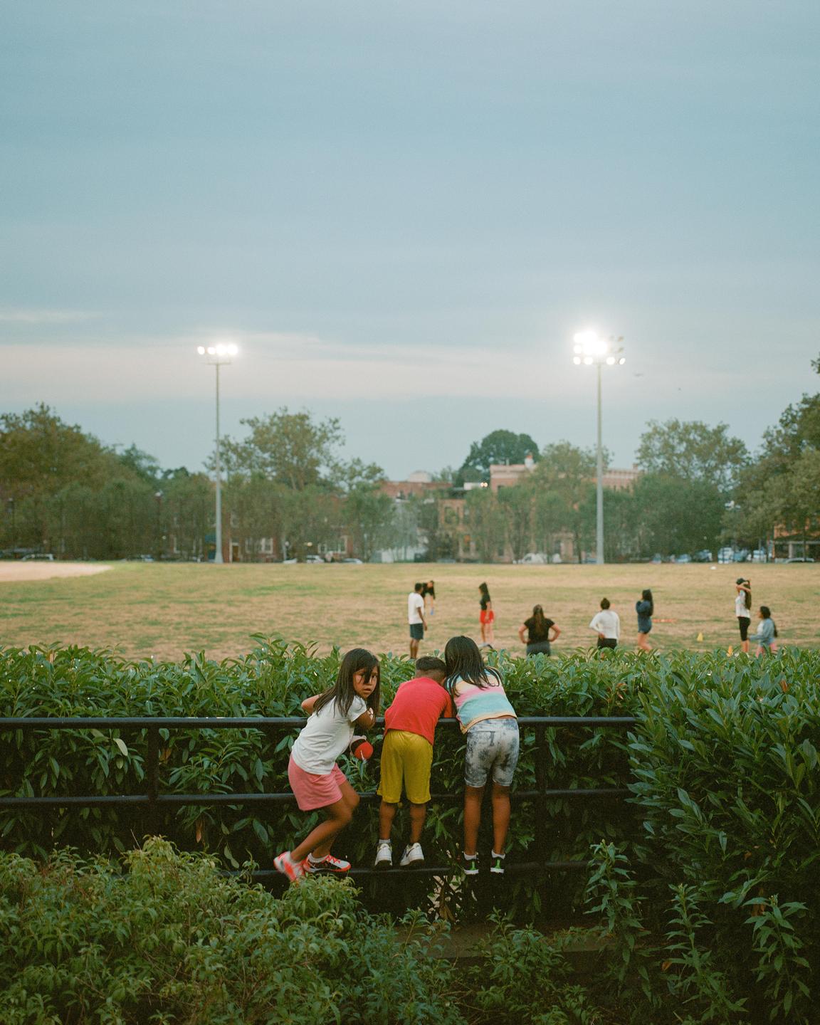 Three children climb a fence to watch people playing on a field under stadium lights.
