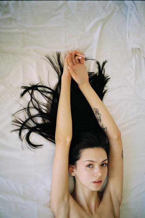 A woman with long dark hair lies on a white bed with her arms stretched above her head.