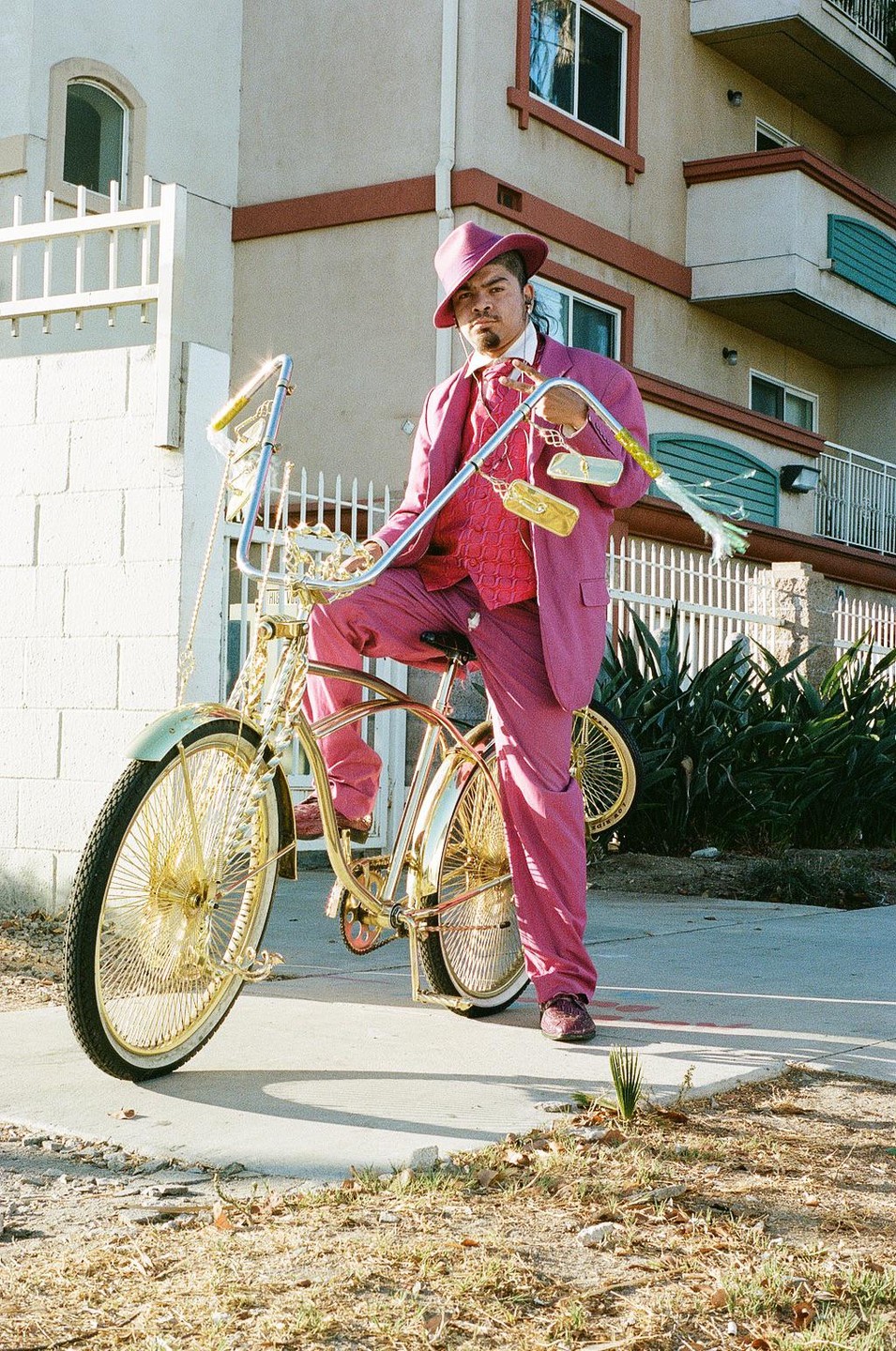 A man in a pink suit and hat sits on a gold lowrider bicycle on a sidewalk.