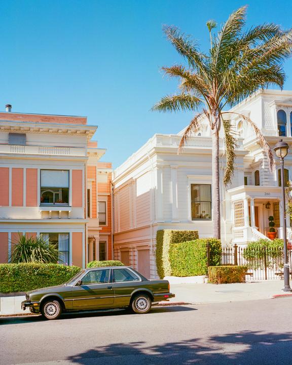 A green car is parked on a street in front of two houses and a palm tree.