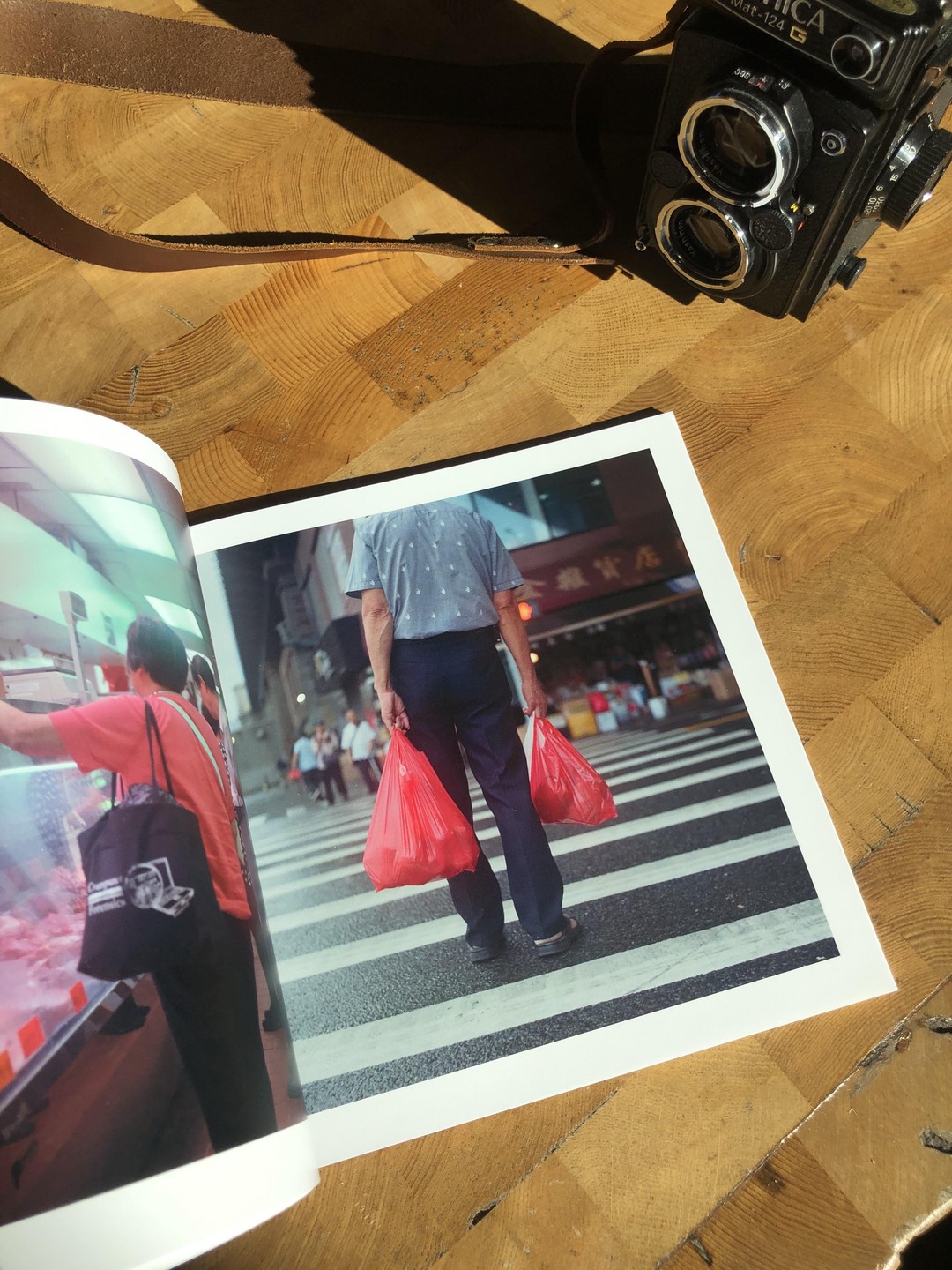 A book is open to a page showing a man carrying two red plastic bags while standing on a crosswalk, next to a camera on a wooden surface.