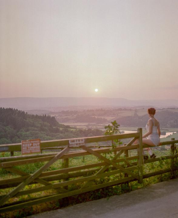 A woman sits on a wooden fence overlooking a valley with a river at sunset.