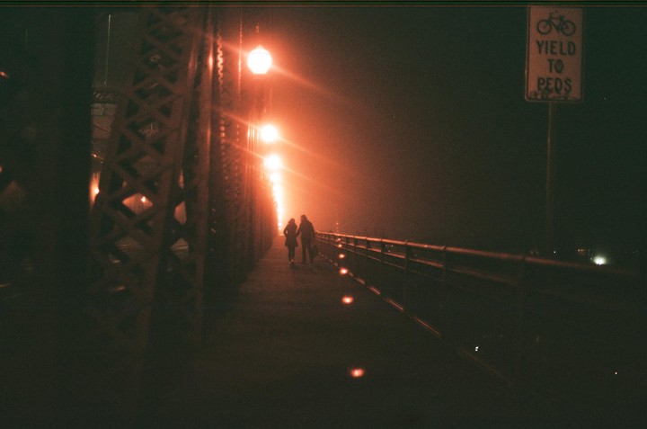 A couple walks hand-in-hand on a bridge at night, illuminated by orange streetlights.