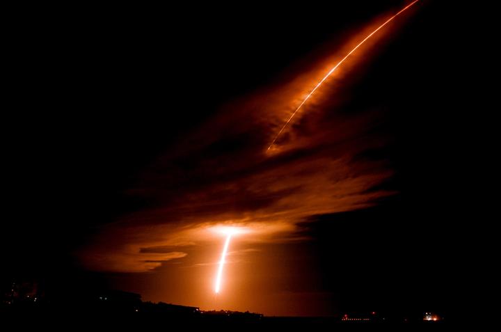 A long exposure photograph captures the fiery ascent of a rocket against a dark night sky, illuminating clouds with an orange glow.