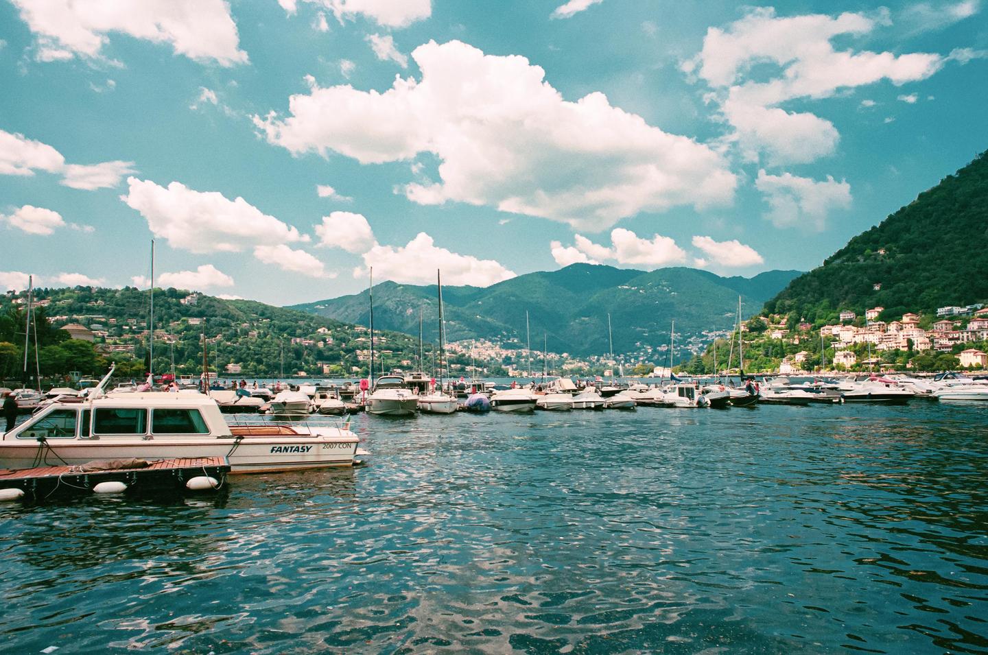 A harbor full of boats is nestled at the base of a mountain covered in houses and trees, under a bright blue sky with fluffy white clouds.