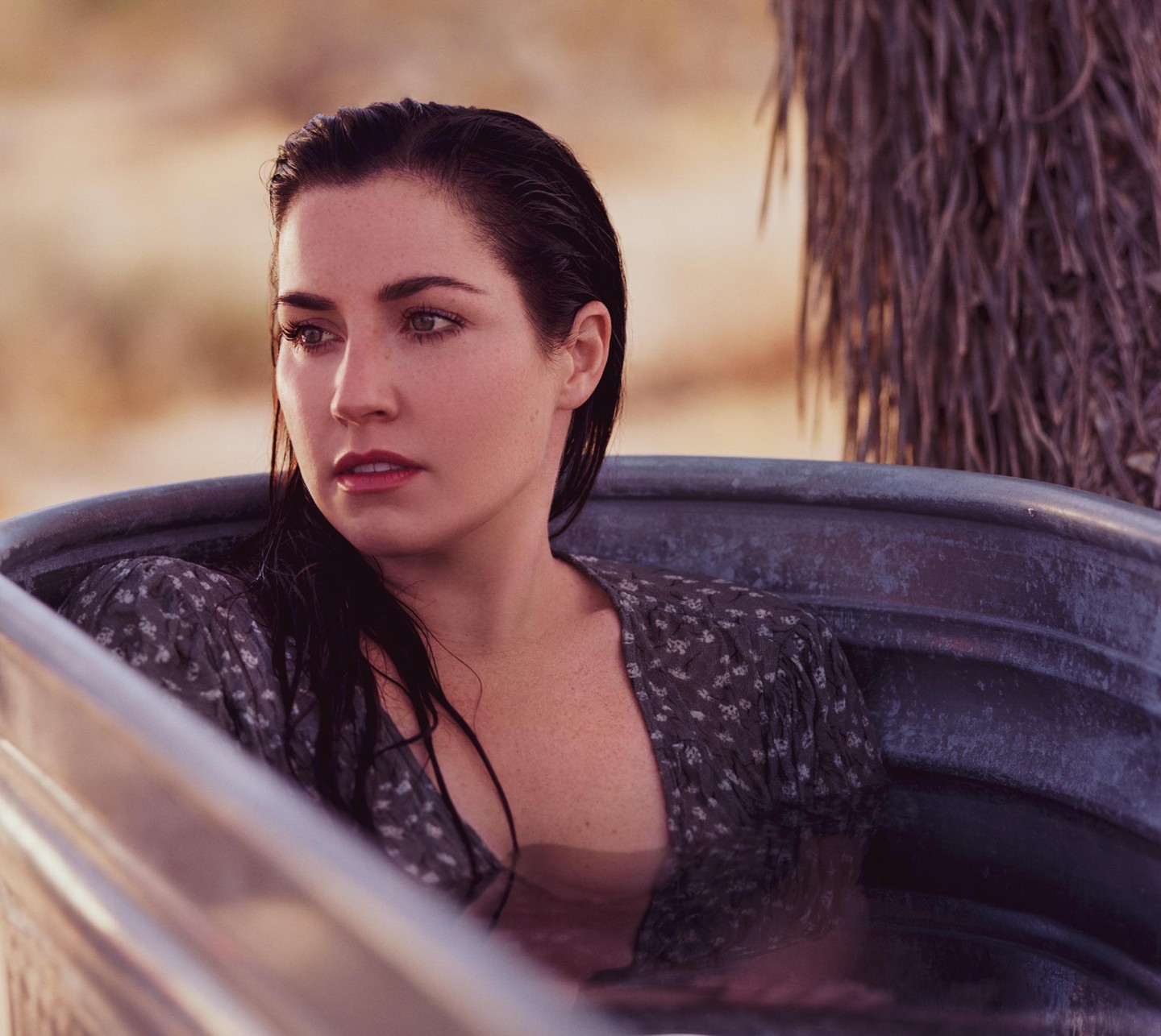 A woman with wet hair is sitting in a metal tub outdoors.