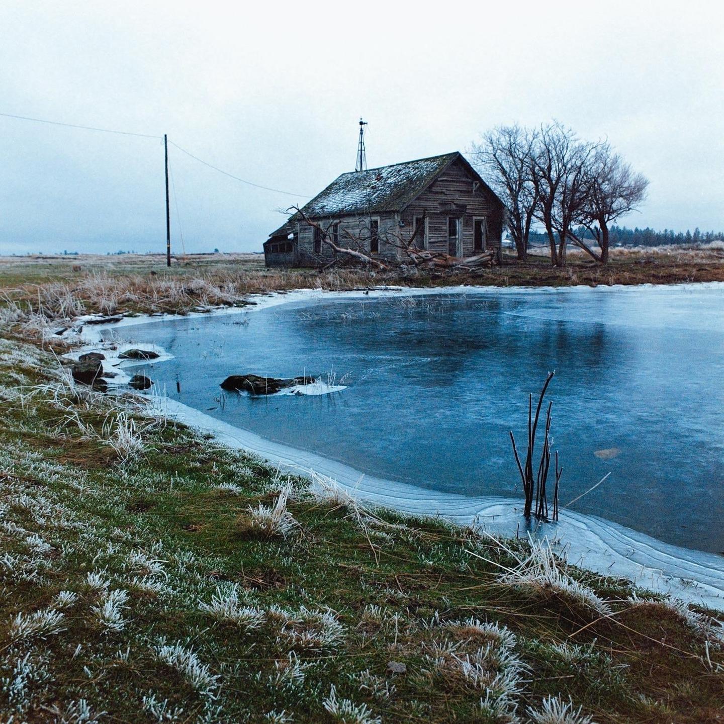 An abandoned wooden house with a windmill on its roof stands beside a partially frozen pond with frosted grass in the foreground.