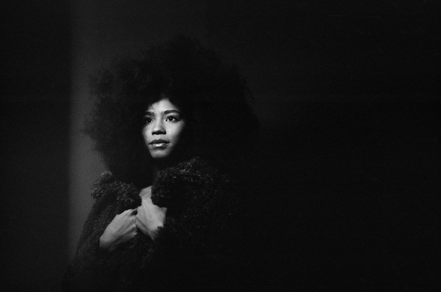 A woman with an afro hairstyle and a textured coat looks up and to the right in a black and white portrait.