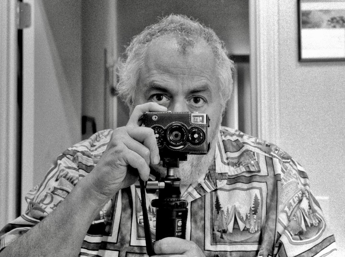 A man with a beard and patterned shirt holds a camera on a tripod in front of his face, taking a self-portrait in a mirror.