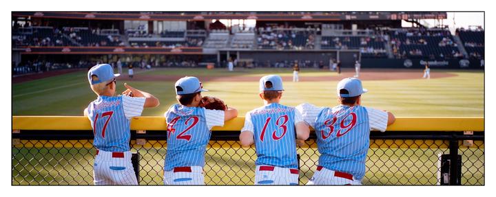 Four boys in baseball uniforms watch a baseball game from behind a fence.
