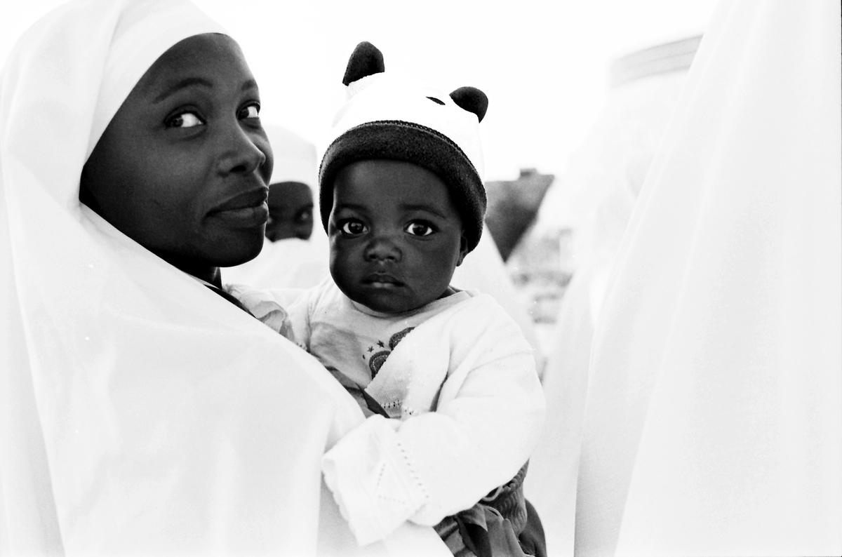 A woman wearing a head covering holds a baby in a hat with ears, with another person visible in the background.