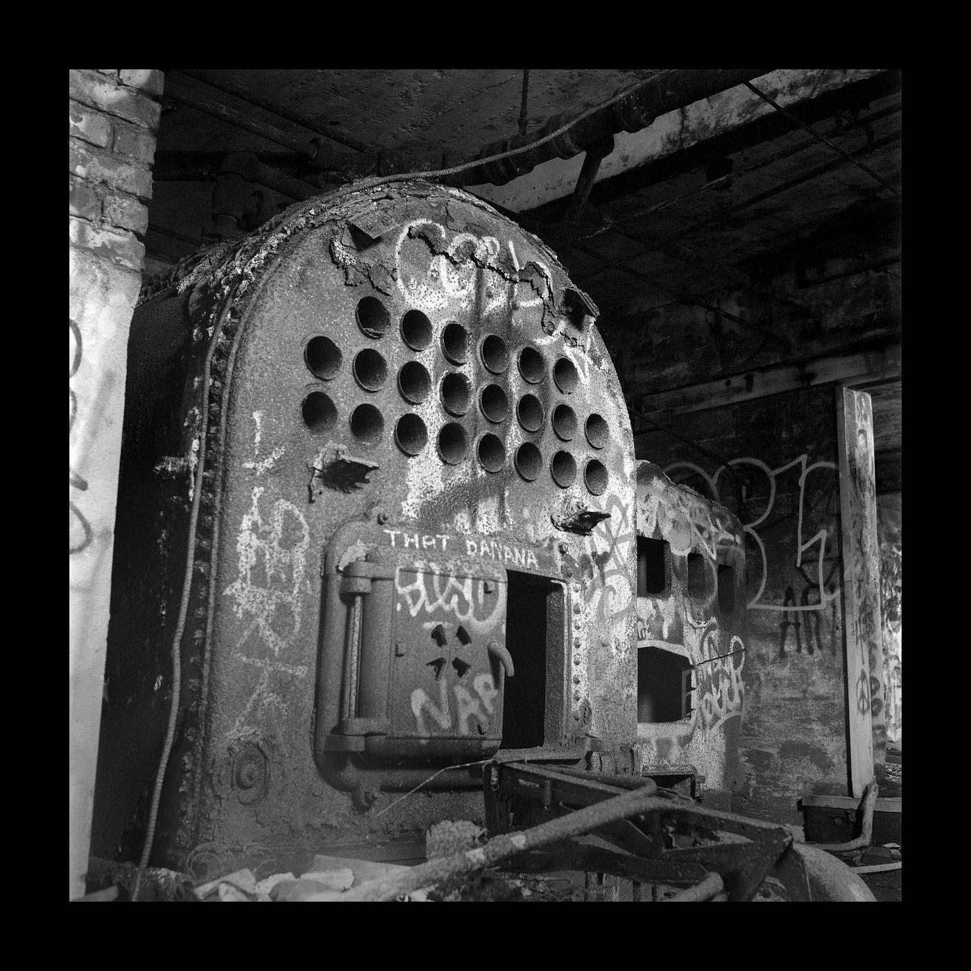 A large, rusty furnace covered in graffiti stands in a dark, abandoned room.