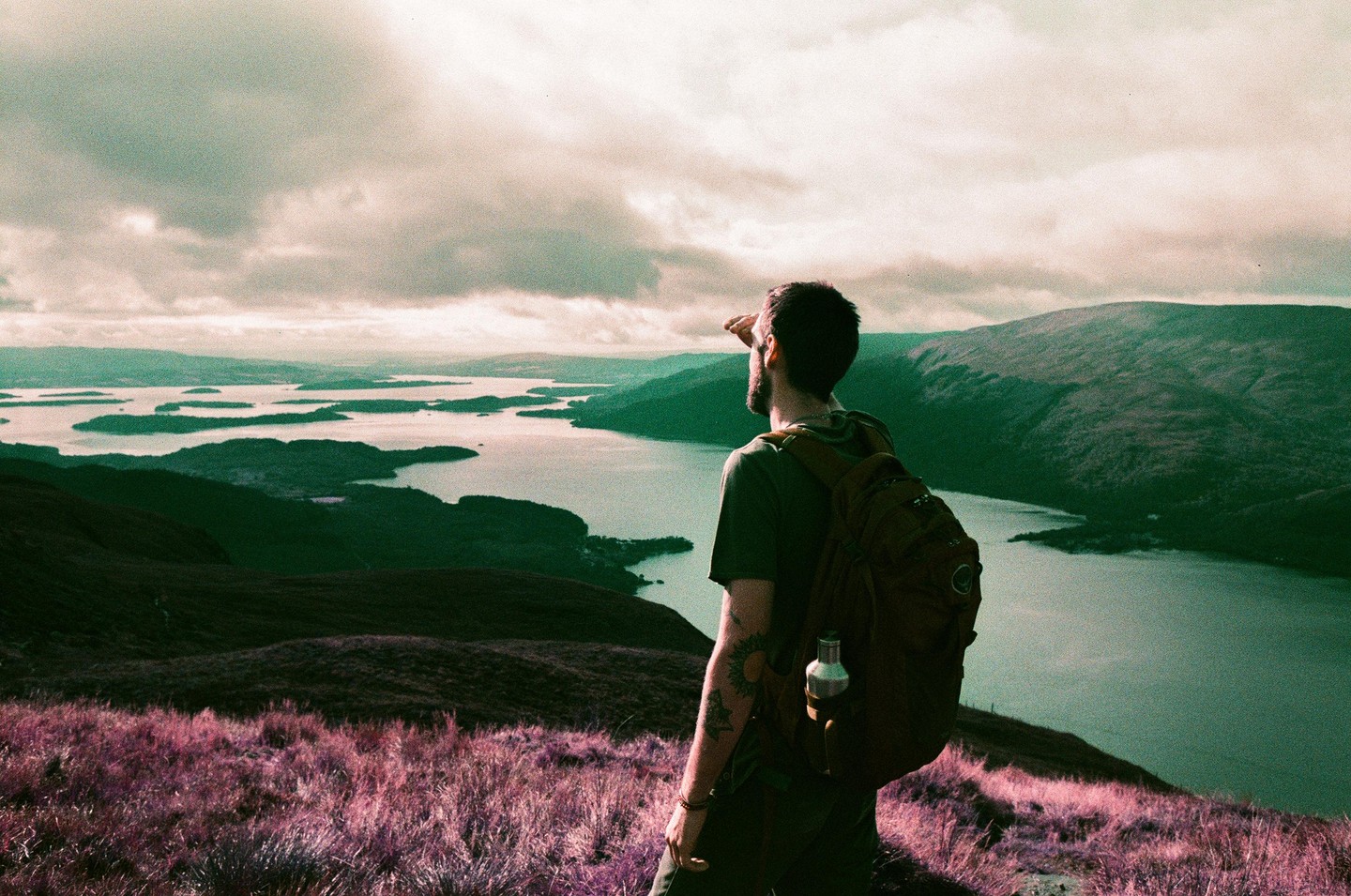A man with a backpack stands on a hill overlooking a body of water with many islands.