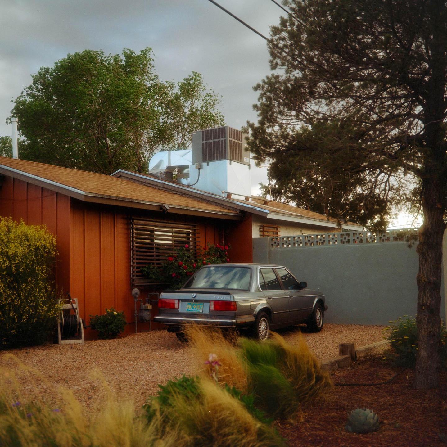 A gray car is parked on a gravel driveway next to a house with a brown roof and orange siding.