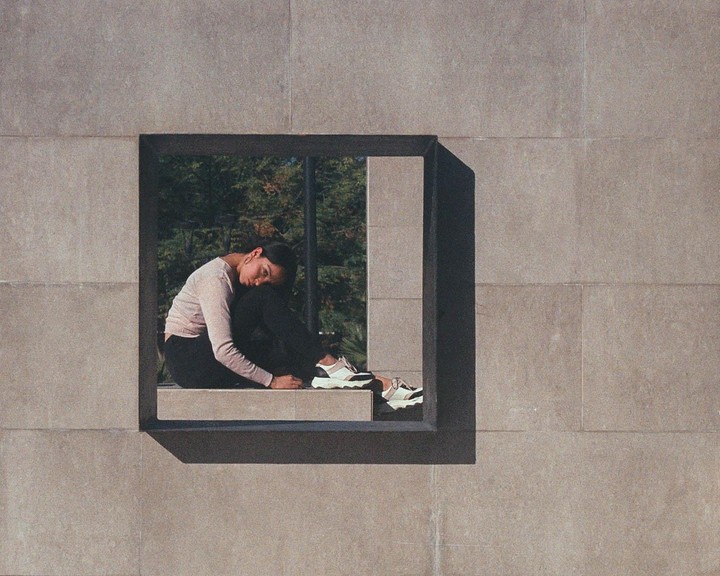 A woman sits curled up in a square window opening of a stone building.