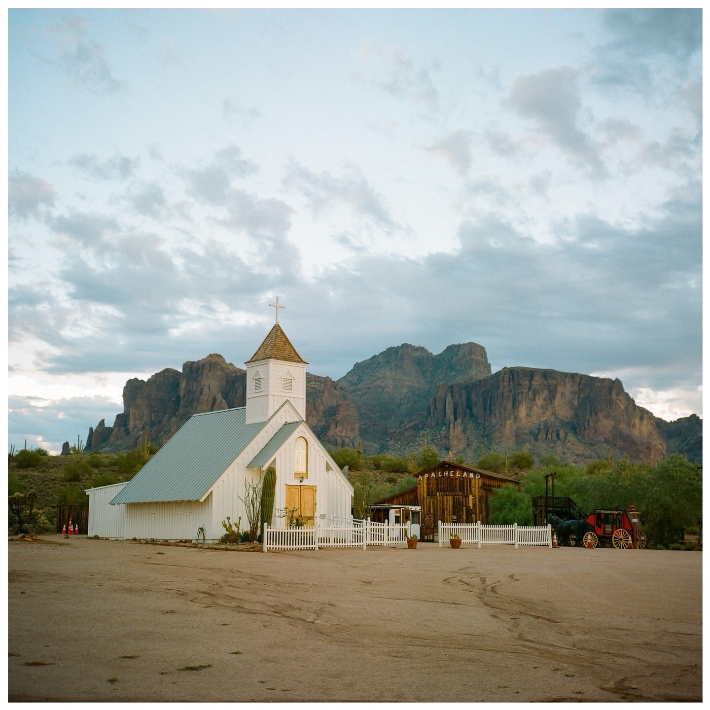 A white church with a steeple stands in a desert landscape with mountains in the background under a cloudy sky.