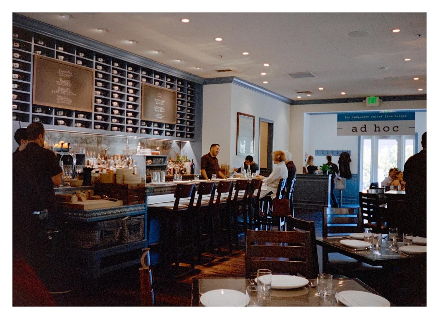 People are seated at a bar and tables in a restaurant with a large wine rack.