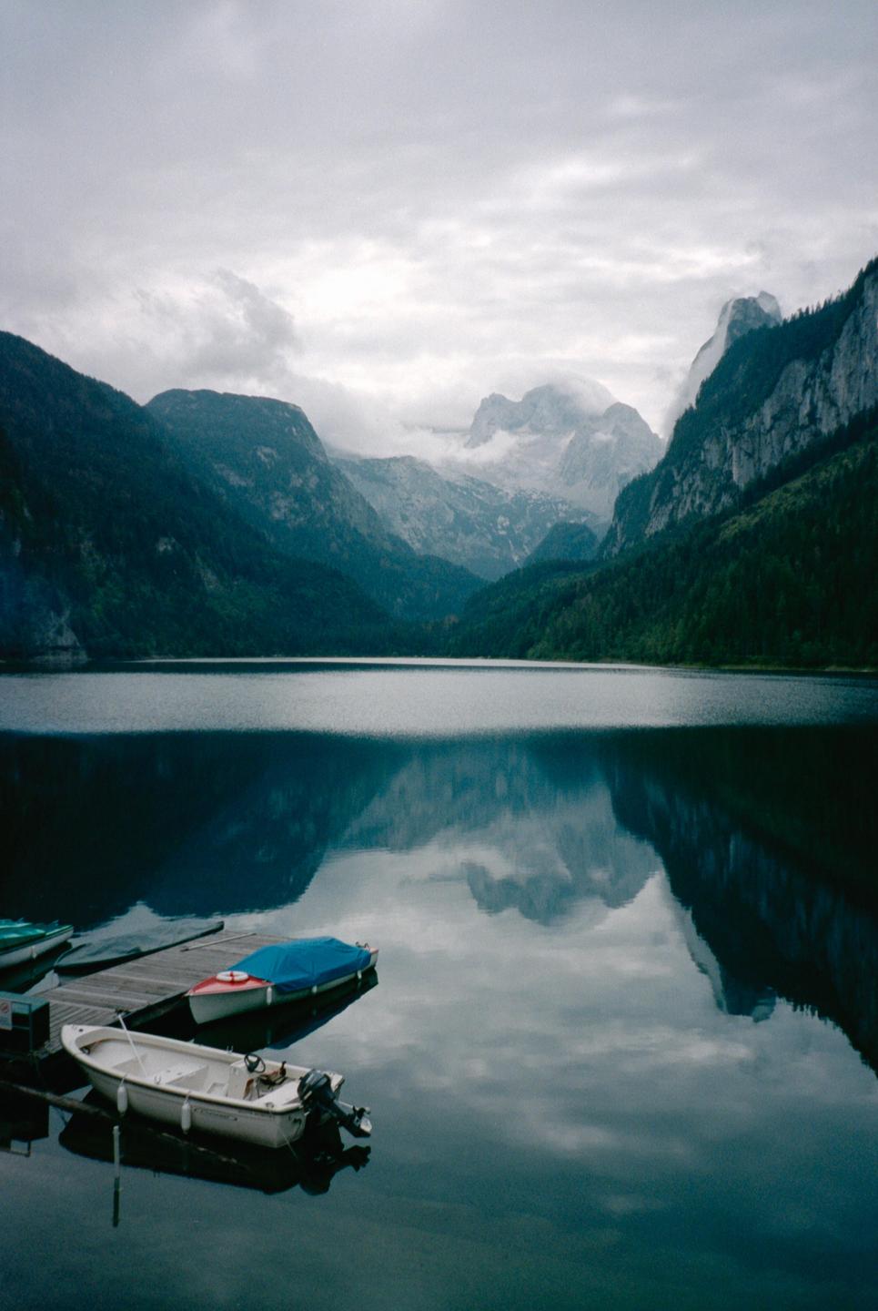 Boats are docked on a calm lake surrounded by mountains under a cloudy sky.
