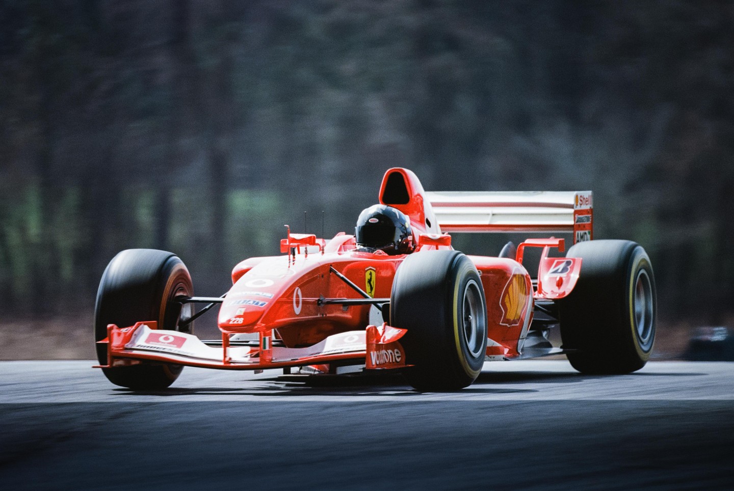 A red formula one race car with a driver in a black helmet speeds down a track with a blurred forest in the background.