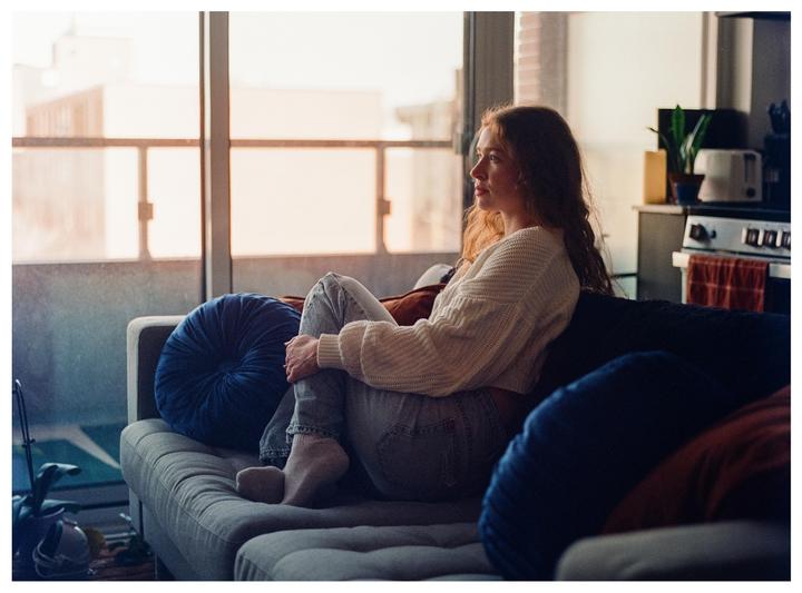 A woman with long hair sits on a sofa in a living room, looking out a window.