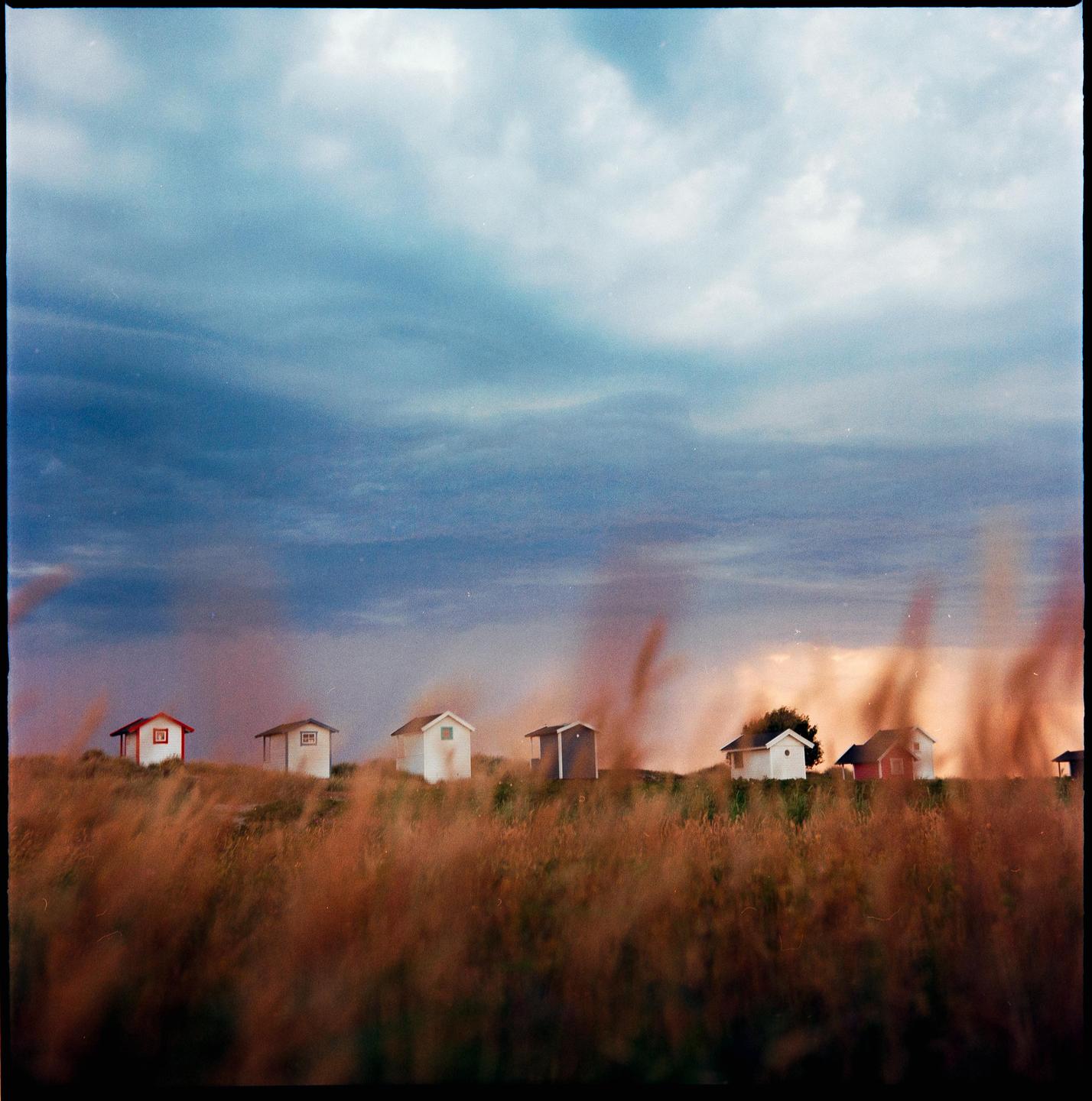 A row of small beach huts sits on a grassy hill under a stormy sunset sky.