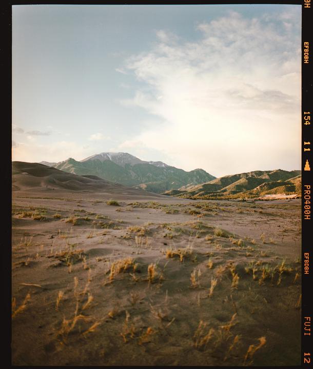 A vast desert landscape with sand dunes and mountains in the background under a partly cloudy sky.