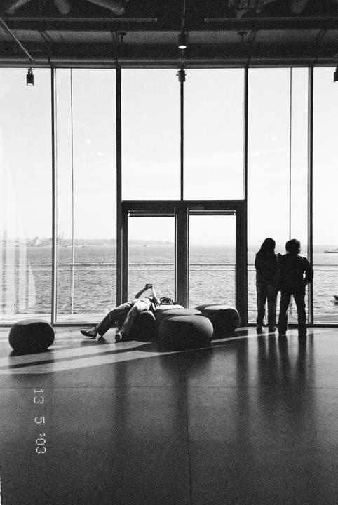 A person reclines on a beanbag while two people stand by a large window overlooking the water.