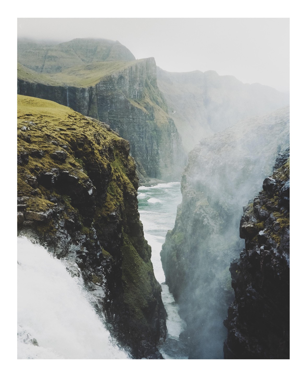 A waterfall cascades into a narrow gorge with a river flowing between steep, rocky cliffs under a hazy sky.