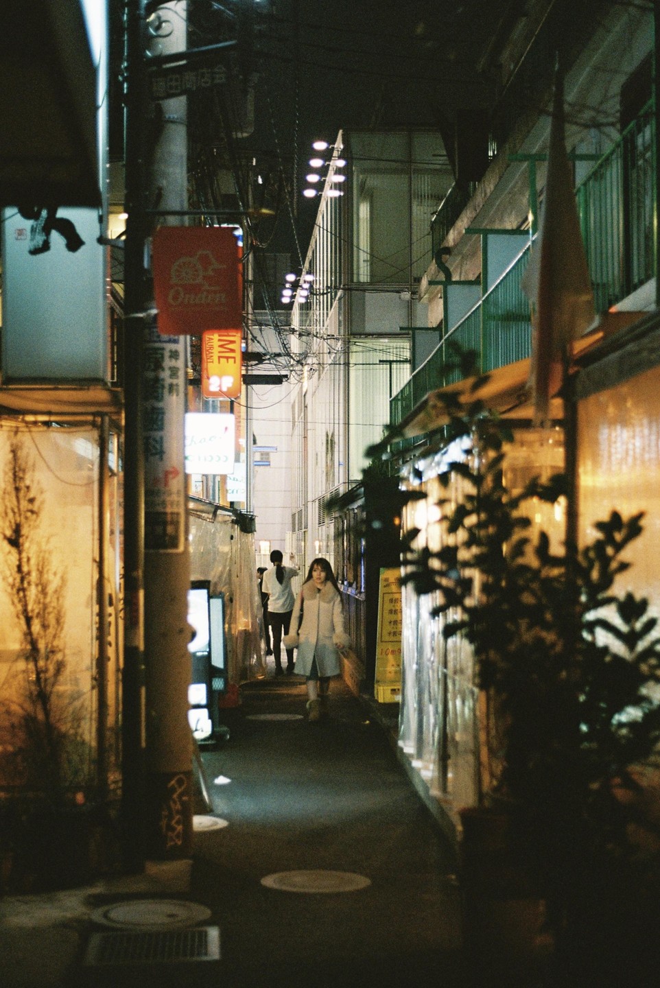 A woman walks down a narrow alley at night with glowing storefronts and signs.