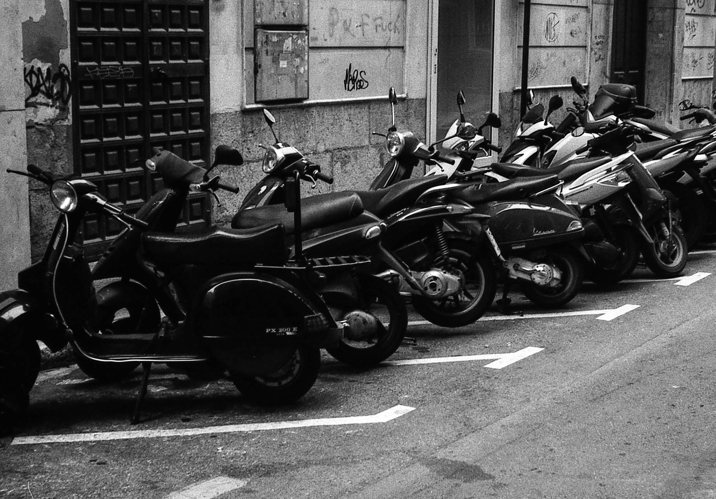 A line of scooters is parked on a street next to a building with graffiti.