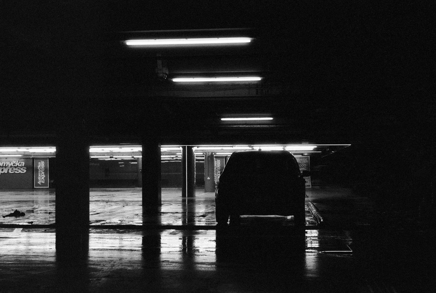 A car is parked in a dark, wet underground parking garage with fluorescent lights.