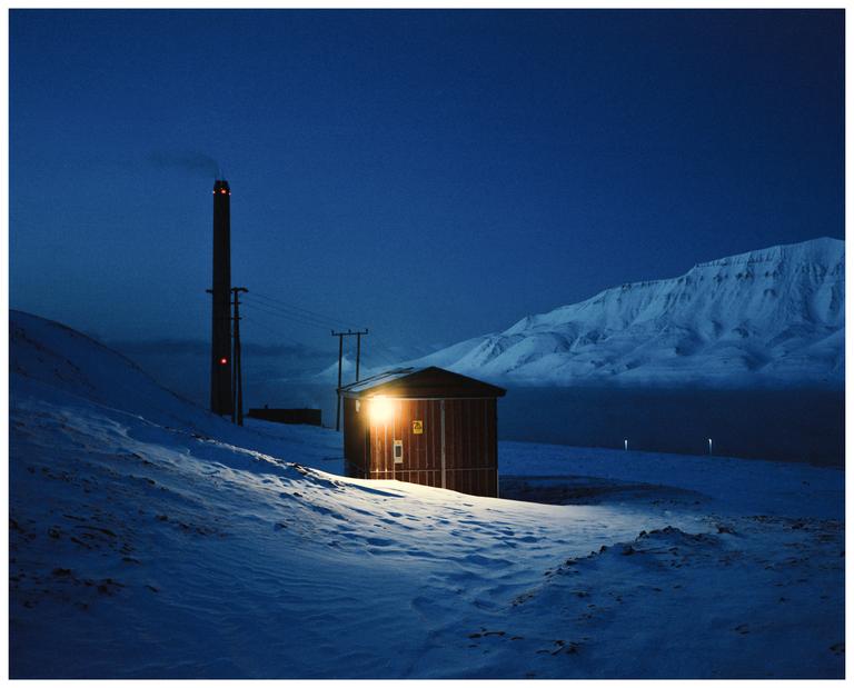A small building with an illuminated window stands in a snow-covered landscape with a tall chimney and mountains in the background at night.