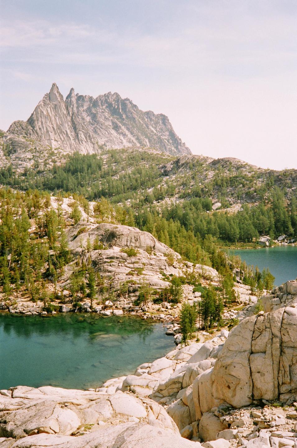 A mountain range with a jagged peak rises above a forest and two clear blue lakes.