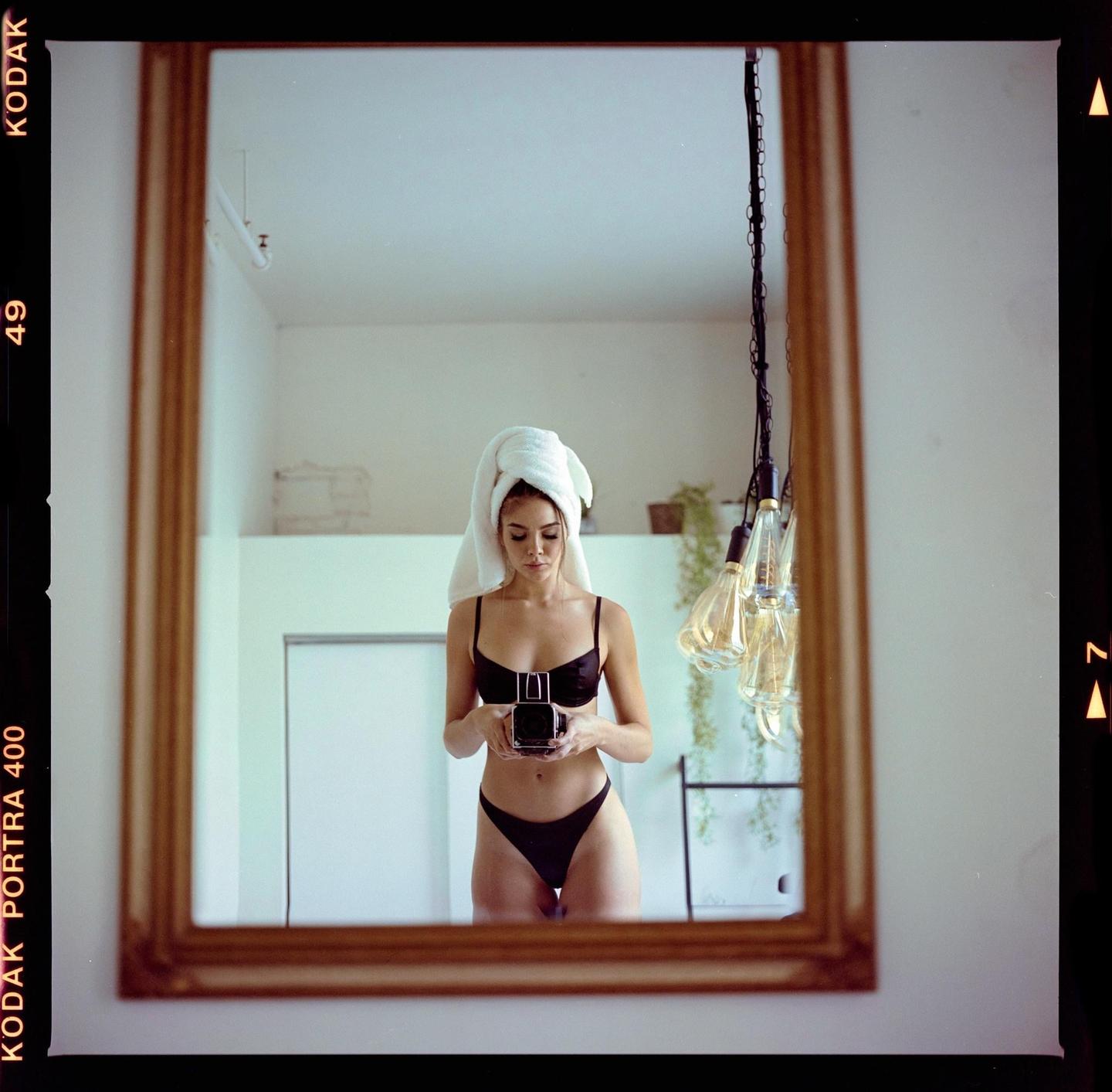 A woman wearing a towel on her head and a black bikini takes a mirror self-portrait with a camera.