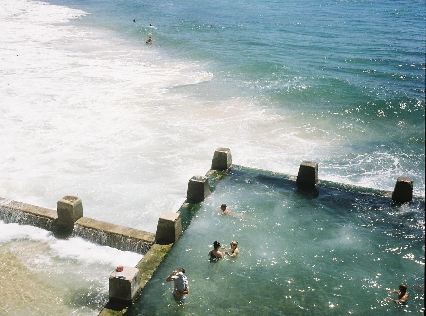 A concrete ocean pool with several people swimming in it is next to a foamy ocean.