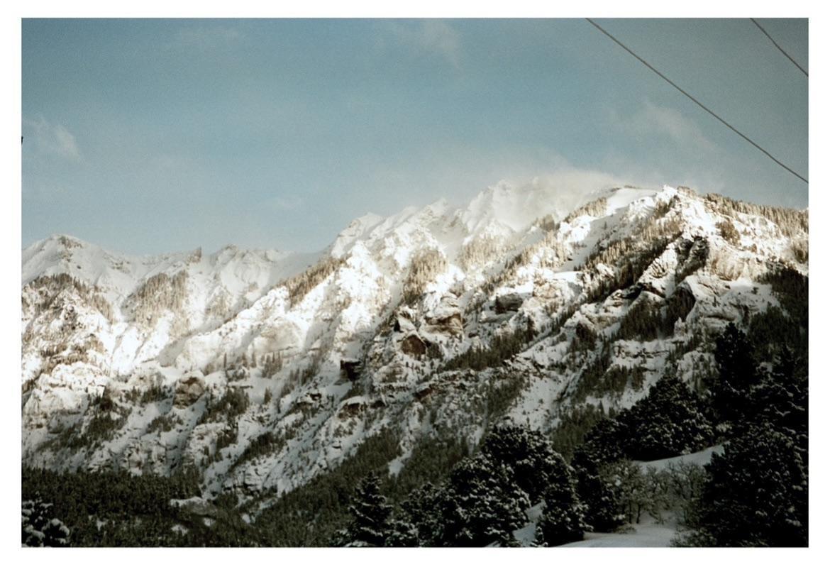 Snow-covered mountains rise under a clear sky, with evergreen trees visible in the foreground.