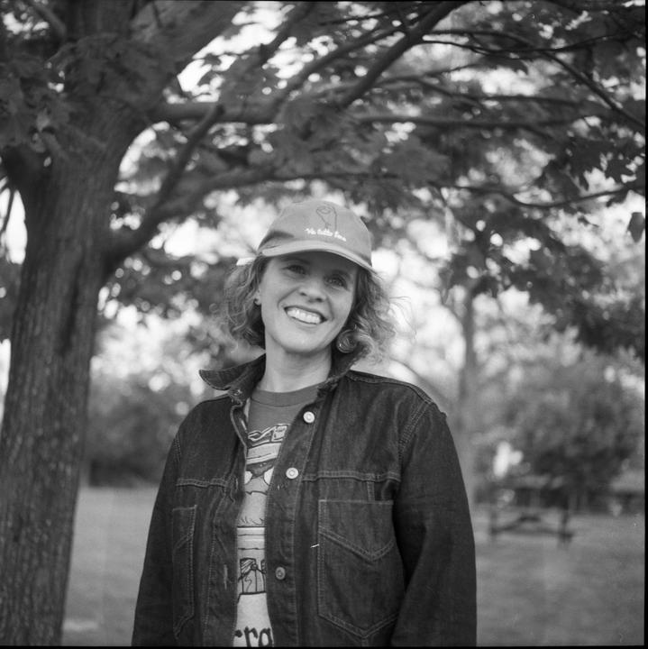 A woman wearing a baseball cap and a denim jacket smiles at the viewer under a tree.