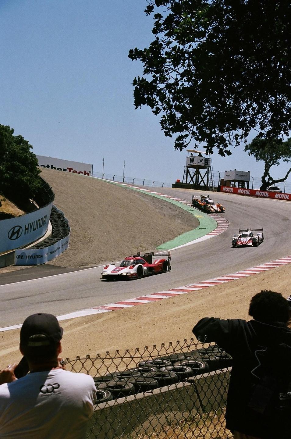 Three race cars speed around a track while two men watch from behind a fence.