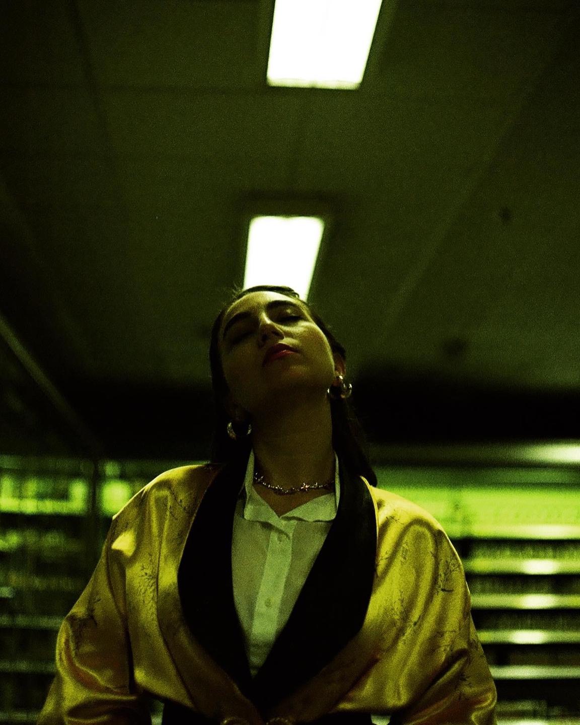 A woman with closed eyes and a gold robe stands under fluorescent lights.