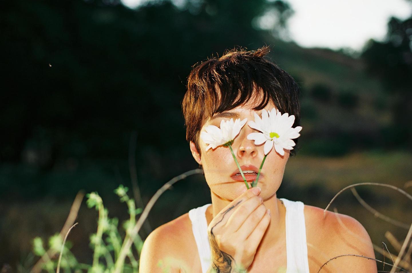 A woman with short hair holds two white daisies over her eyes.