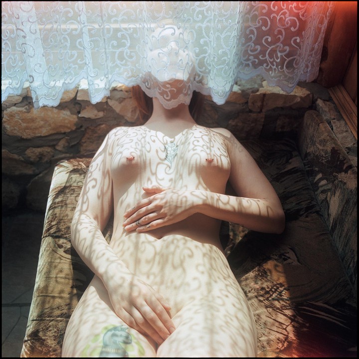 A woman lies nude on a couch with a lace curtain covering her face and casting shadows on her body.