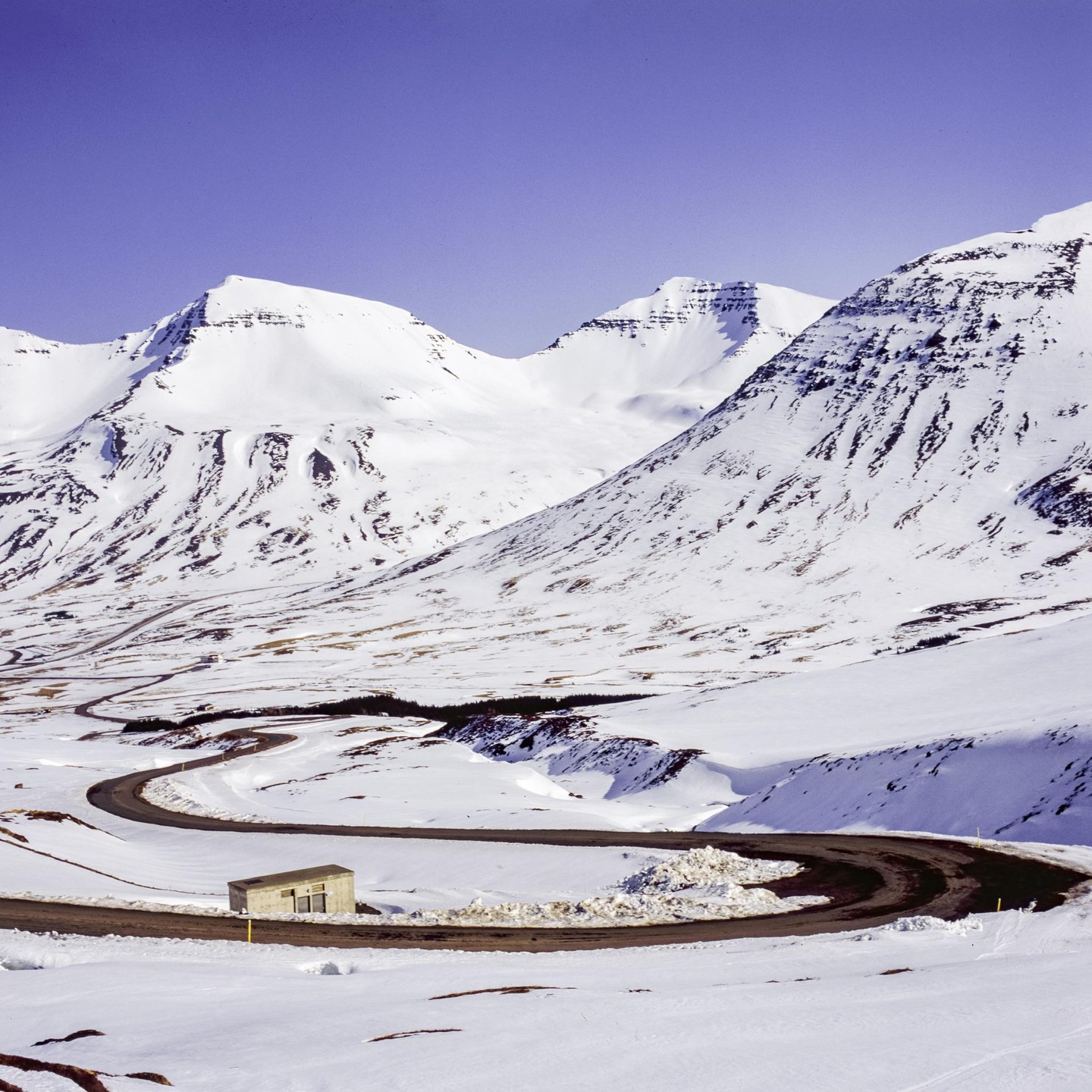 A winding road leads through a snow-covered mountain landscape with a small building beside the road.
