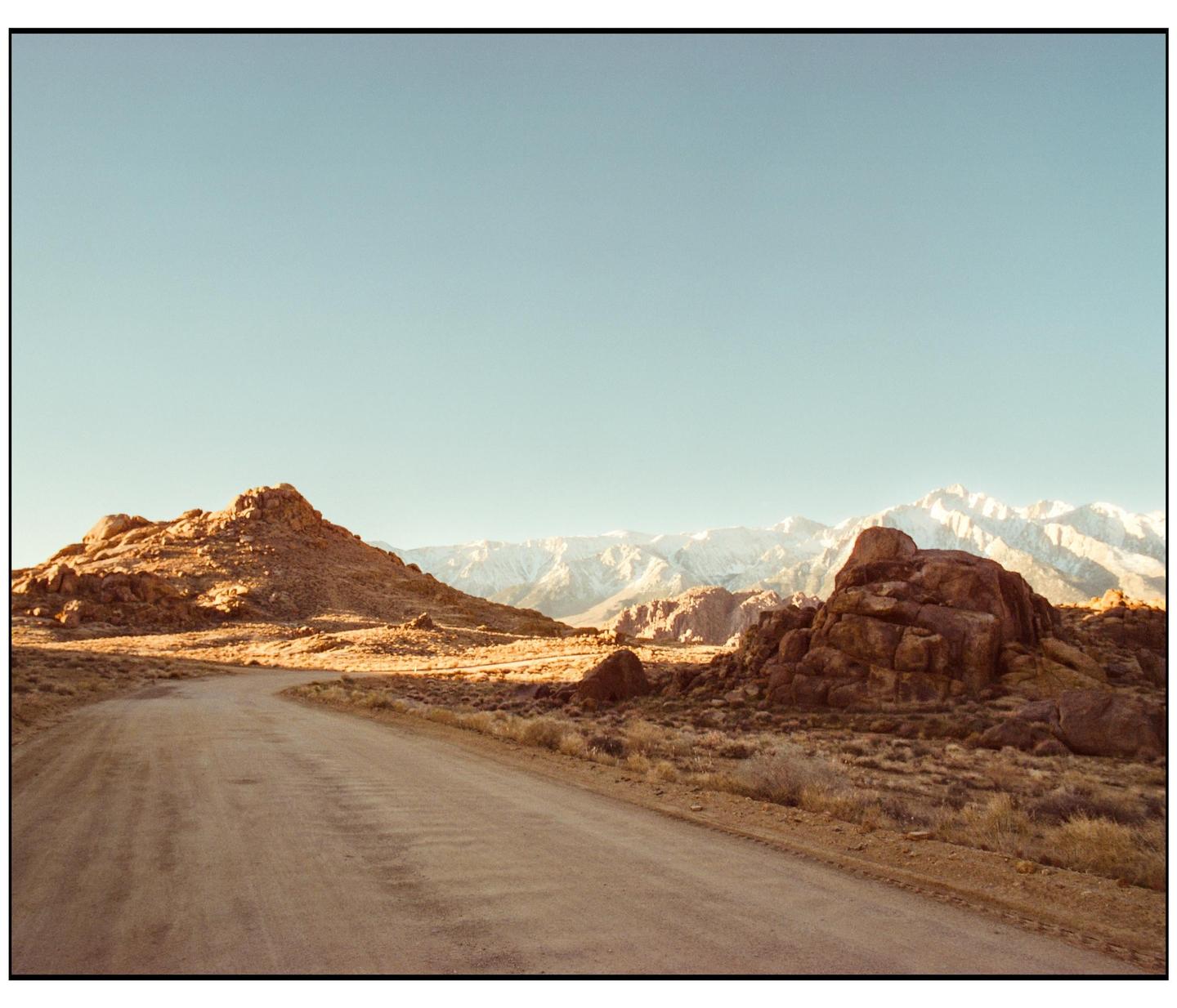 A dirt road winds through a rocky, arid landscape with snow-capped mountains in the distance under a clear sky.