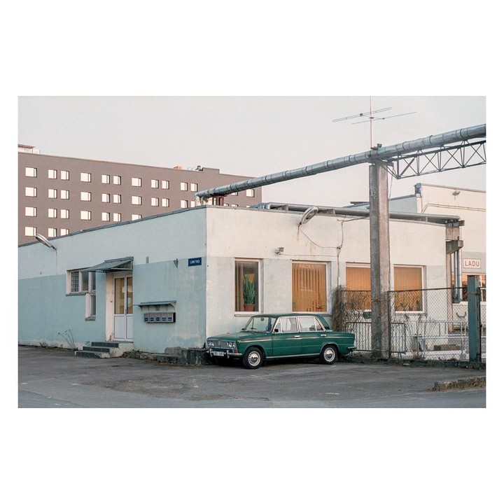 A green car is parked in front of a light blue building with a large pipe running above it.