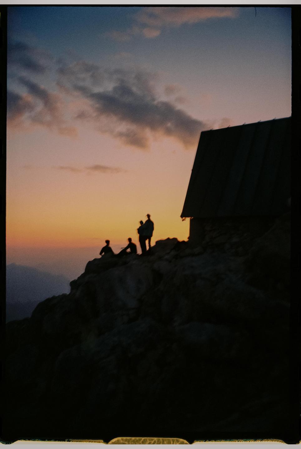 Silhouettes of people and a building are visible on a rocky mountain at sunset.